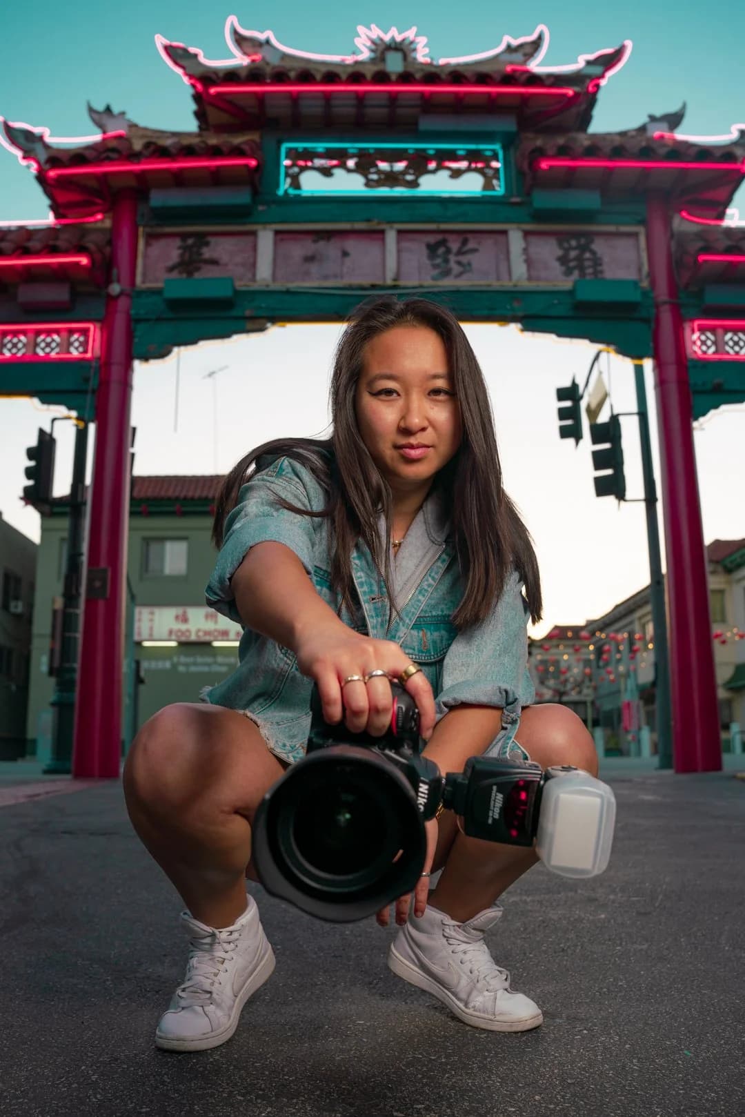 Olivia Wong crouching in front of the neon-lit Chinatown gate in Los Angeles, holding a camera with a long lens toward the viewer