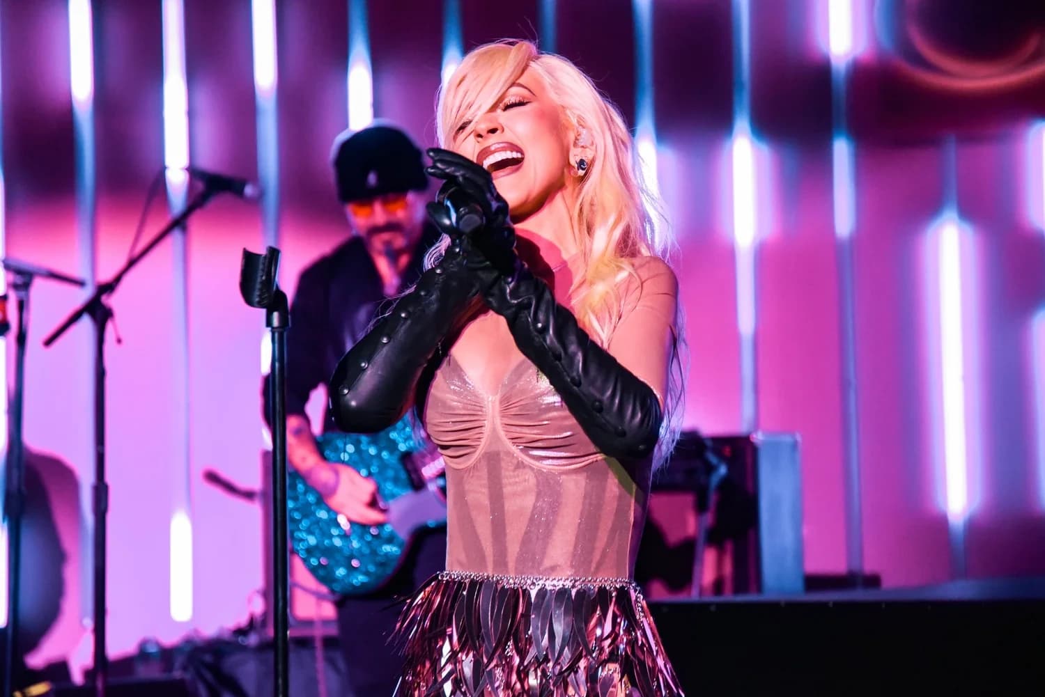 A singer mid-note at the microphone in long black leather gloves and a metallic fringed skirt, backed by vertical light tubes in a pink stage wash
