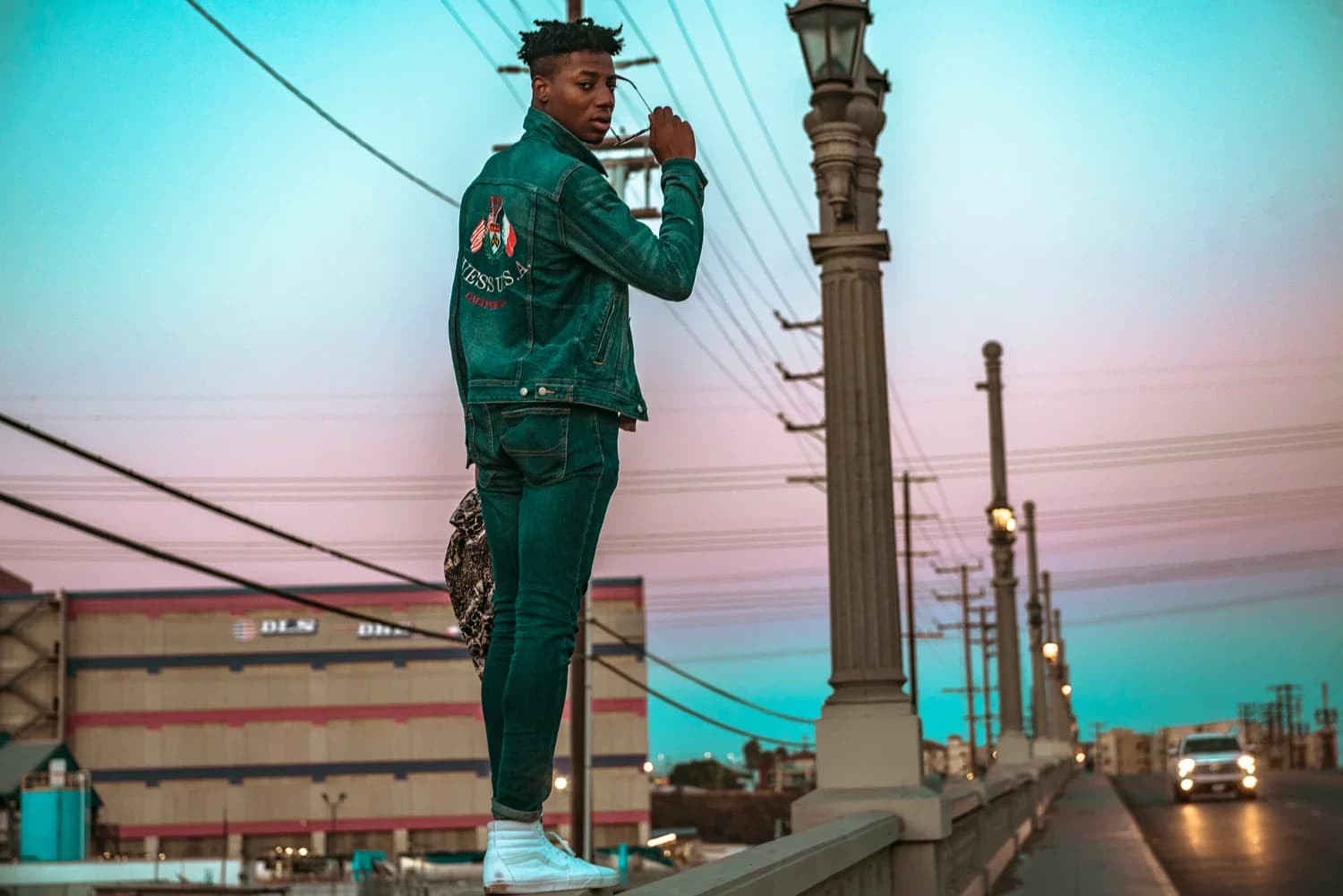 A man in a green denim jacket and jeans standing on a bridge parapet at dusk, looking back over his shoulder as he lowers his sunglasses, the sky behind him graded teal into pink