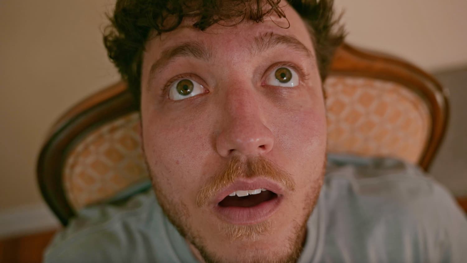 An extreme low-angle close-up of a man with wide eyes and an open mouth, lying back against an upholstered headboard — a still from the music video Angry Mob