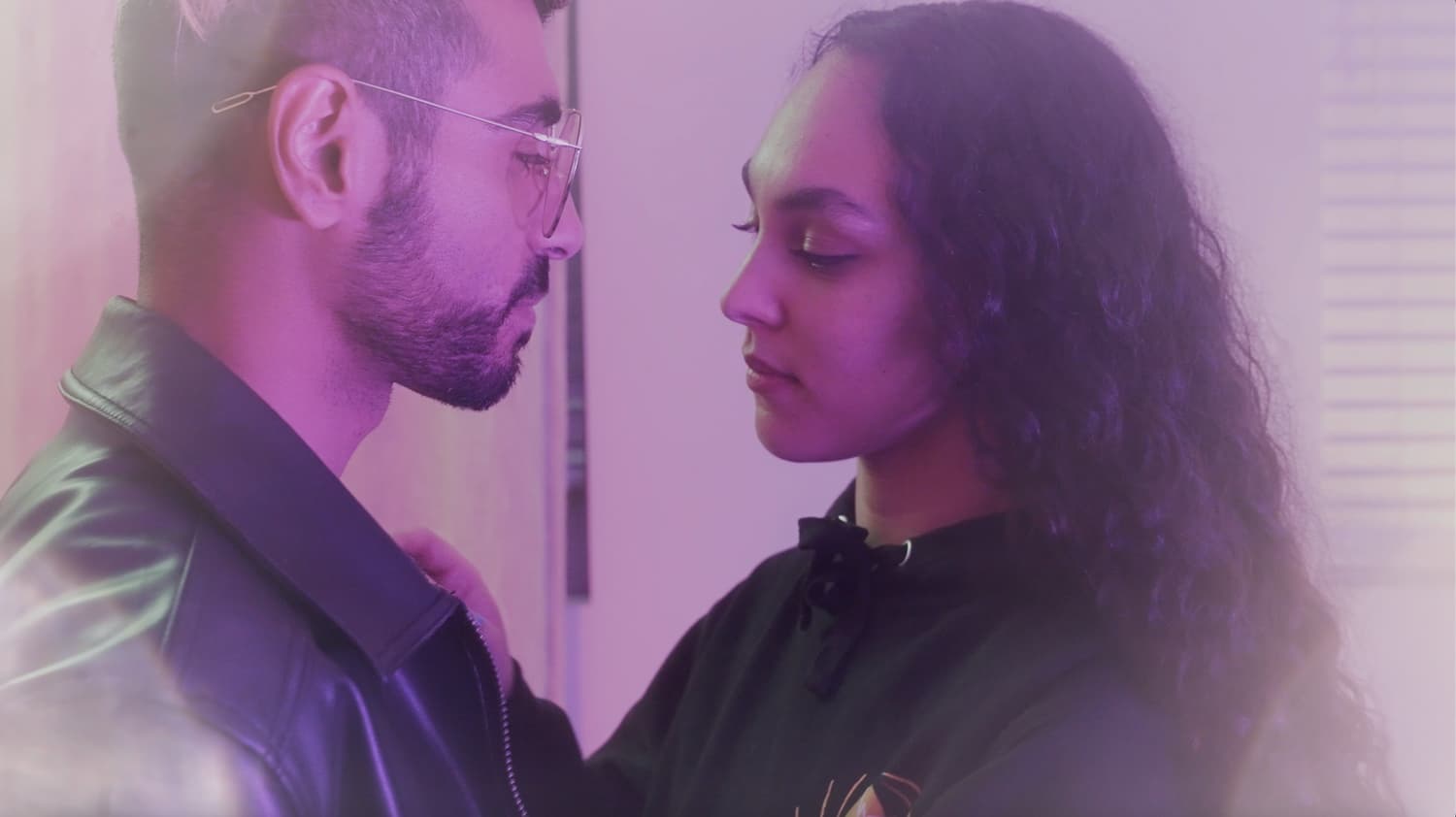 A man in a leather jacket and a woman with long curly hair stand face to face in pink-purple light, her hand at his collar — a still from the music video Fantasy