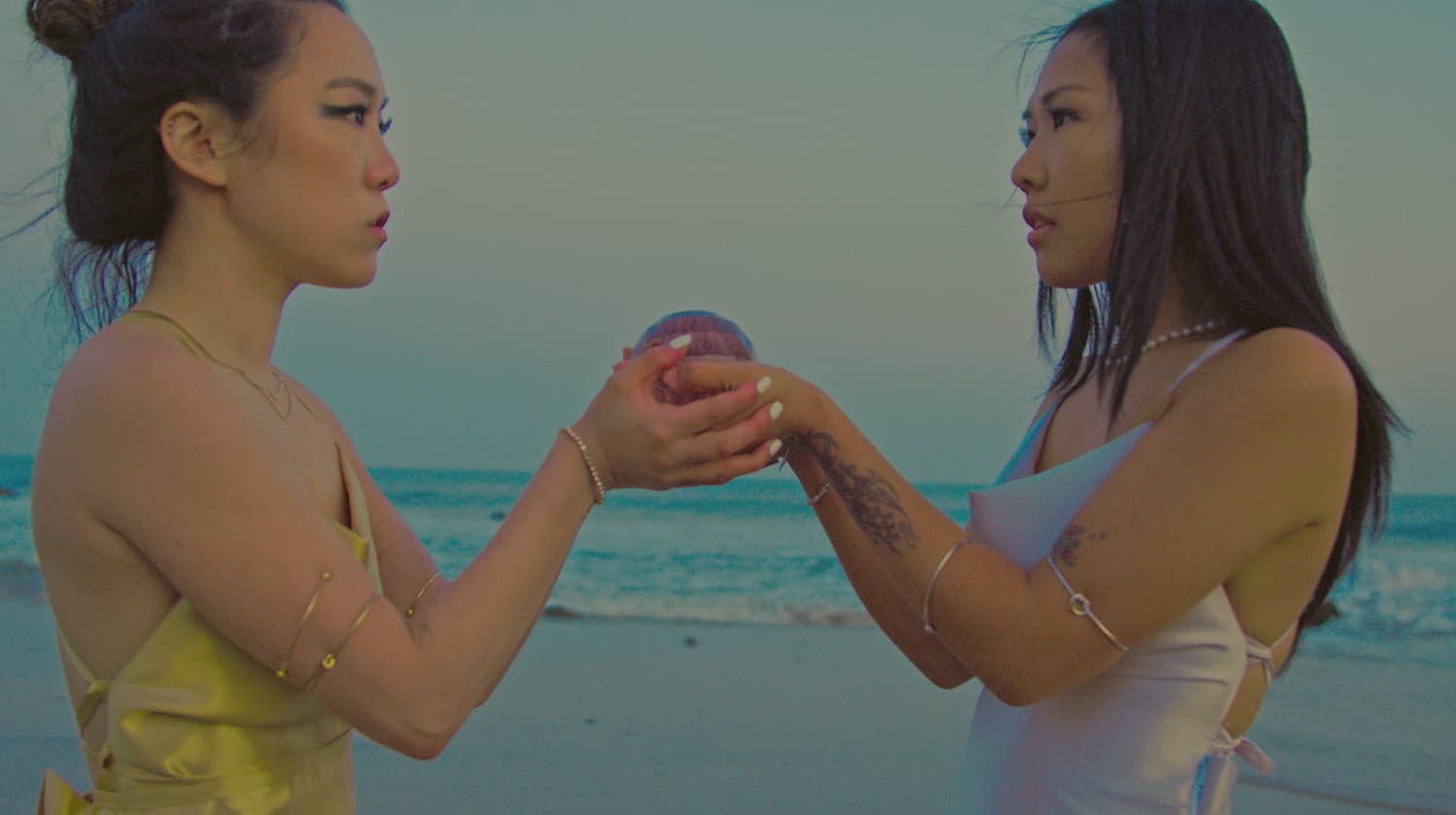Two women face each other on a beach at dusk, cupping a small iridescent sphere between their hands — a still from Merge