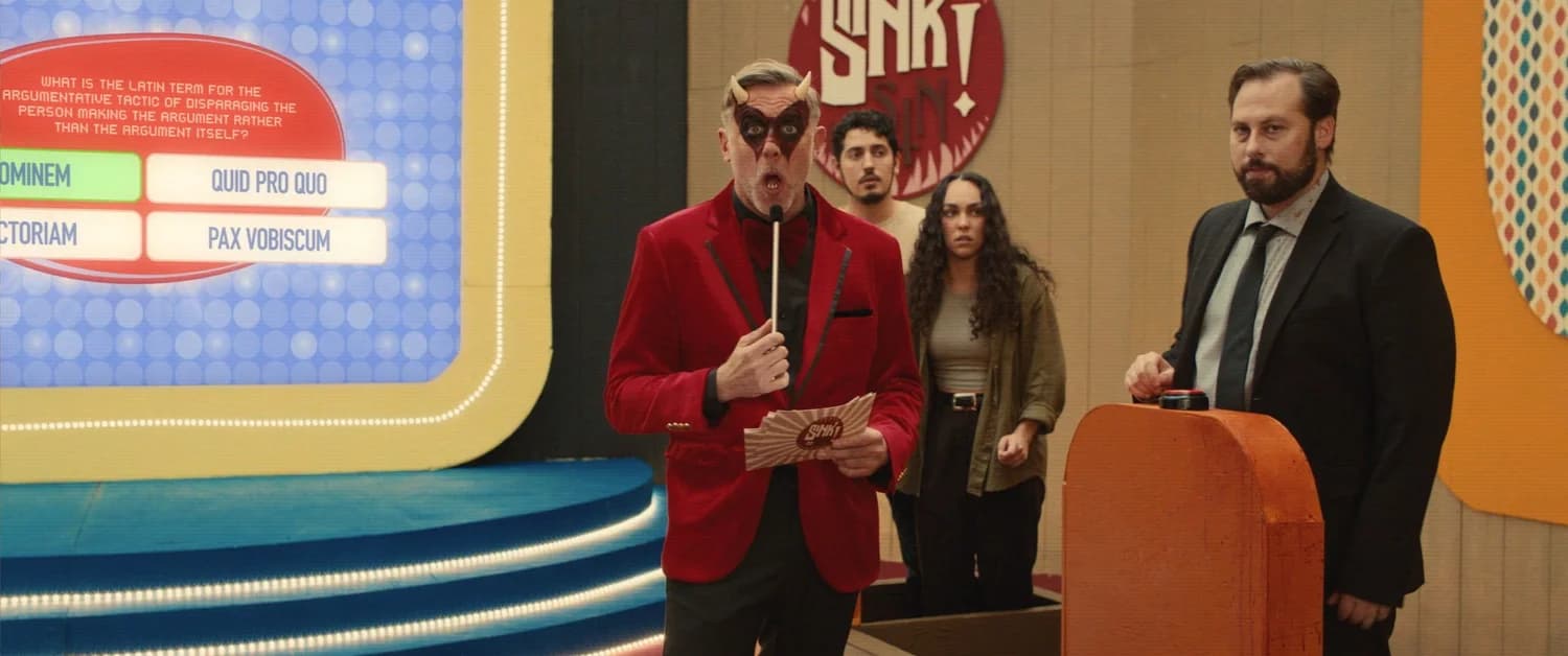 A gameshow host in a red blazer and devil horns holds a microphone on a brightly lit set, two contestants behind him and a trivia question on the screen beside — a still from Sink or Sin
