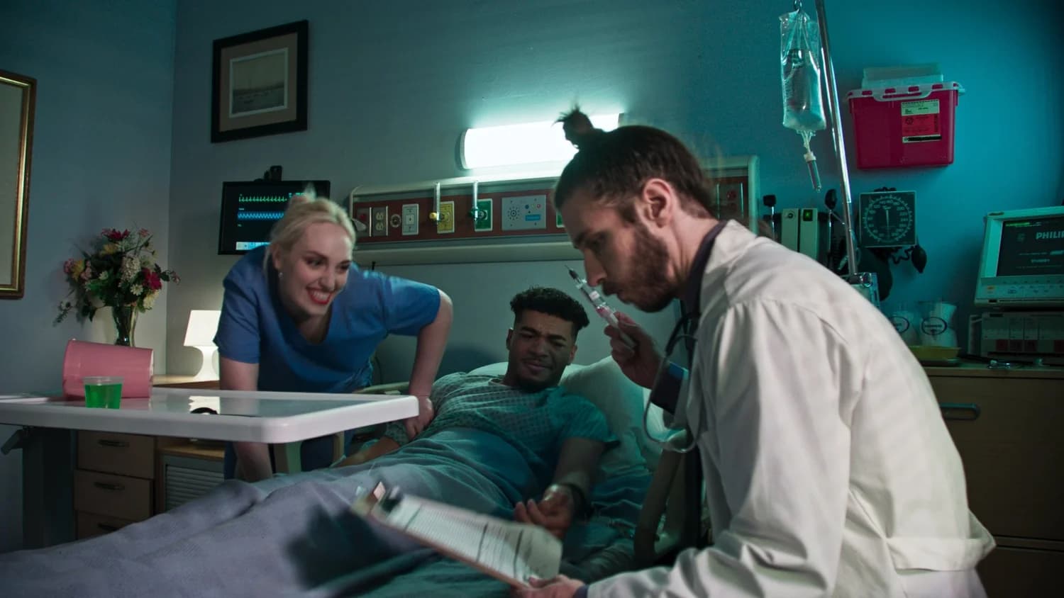 A hospital room lit teal — a nurse leans over a patient in bed while a doctor beside them holds up a syringe — a still from the music video Solo